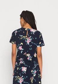 Blusa floral azul-marinho com mangas curtas, apresentando padrões de flores em rosa e amarelo, uma abertura em forma de chave nas costas e um tecido suave e texturizado.