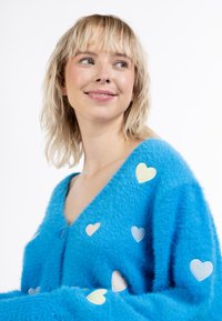 Cardigan blu morbido con scollo a V, decorato con toppe a forma di cuore multicolori di diverse dimensioni sul davanti e sulle maniche, e chiusura con bottoni.
