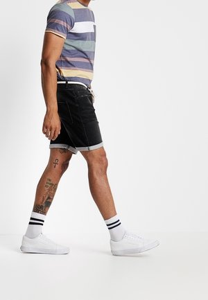 Randig T-shirt i flera färger; svarta jeansshorts med uppvikta fållar; vita sneakers; vita strumpor med svarta ränder; synliga tatueringar.