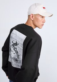 Felpa nera con un grande grafico in bianco e nero sul retro, caratterizzato da immagini astratte. Completata da un cappellino da baseball bianco.