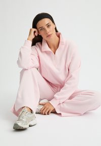 Lichtroze, ritssluiting pullover met kraag, gecombineerd met bijpassende losvallende broek. Zilveren sportschoenen accentueren de outfit. Gladde textuur.