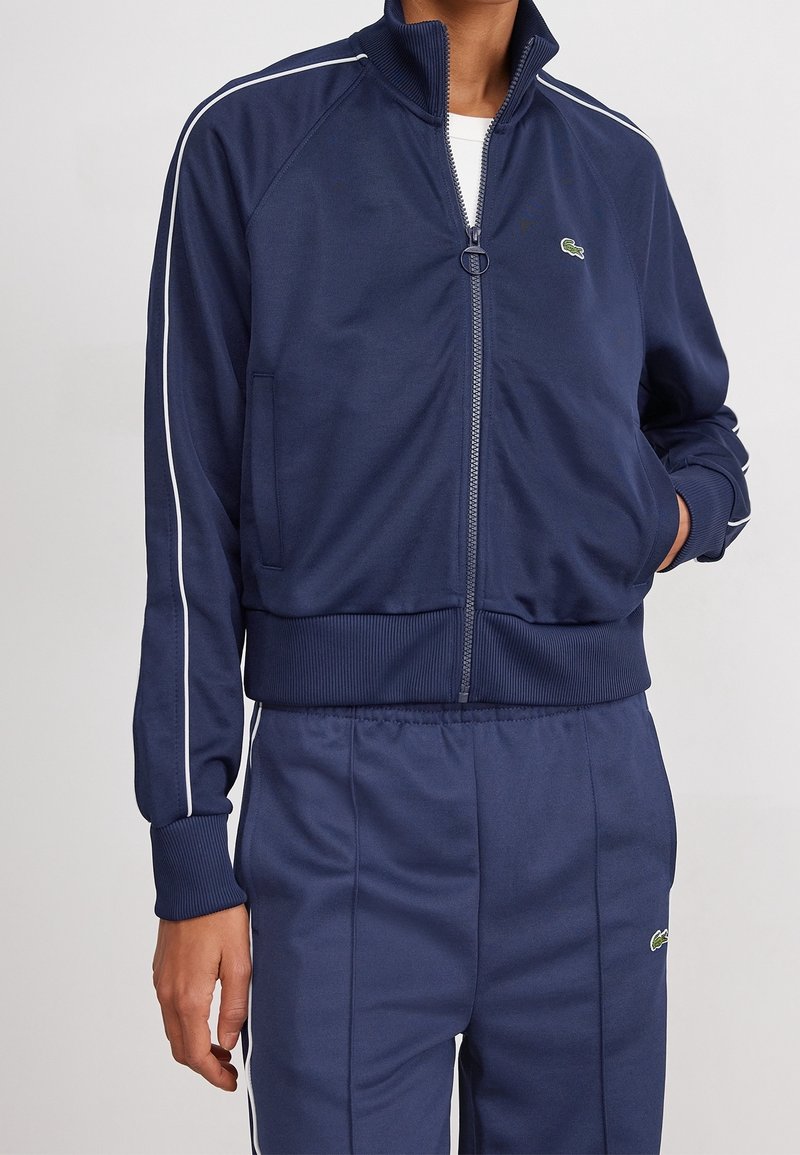 Chaqueta de chándal azul marino con cremallera, puños y dobladillo de canalé, que presenta franjas laterales blancas y un pequeño logo verde en el lado izquierdo del pecho. Tejido suave.