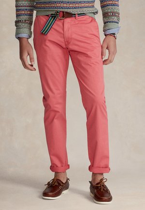 Homme portant un pantalon rose corail retroussé, des chaussures bateau en cuir marron, un pull à motifs, une ceinture rayée et une montre au poignet gauche.