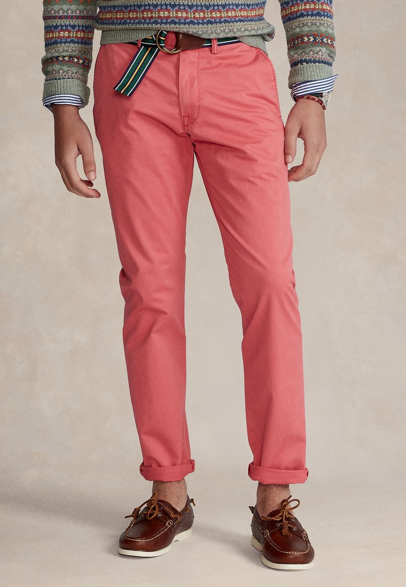 Homme portant un pantalon rose corail retroussé, des chaussures bateau en cuir marron, un pull à motifs, une ceinture rayée et une montre au poignet gauche.