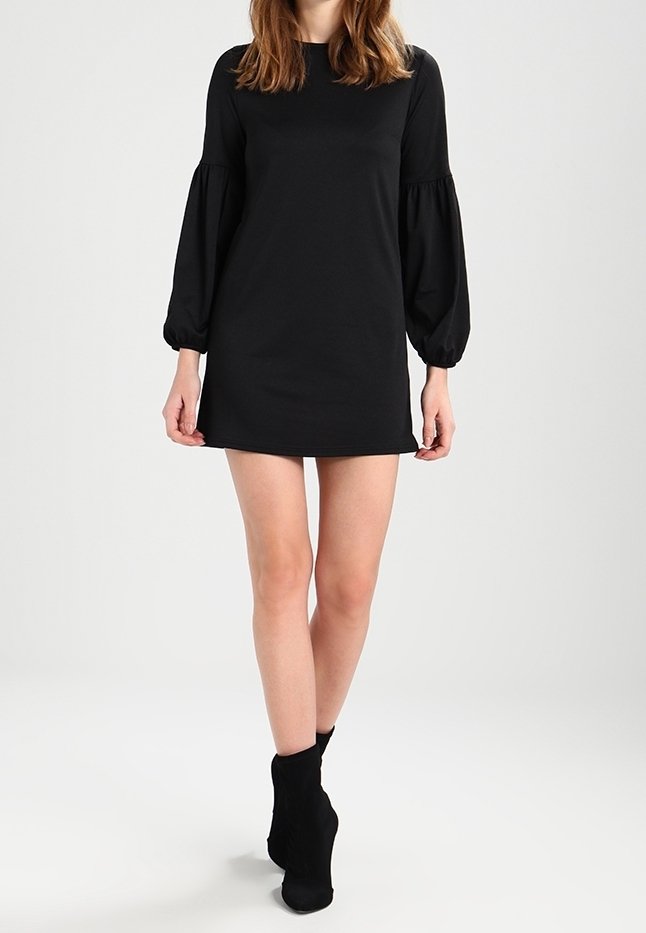 Femme portant une mini-robe noire à manches longues bouffantes et des bottines noires, debout devant un fond clair uni.