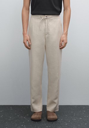 Mand iført beige bukser med snor, sort skjorte og brune sko står på en mørk pletgulv mod en lysegrå væg.