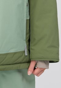 Giacca stratificata verde e verde chiaro con tasca con zip, dotata di etichetta con logo e maniche con polsini per una maggiore calore e comfort.