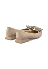 Ballerines en suède beige avec un petit talon carré, présentant un nœud décoratif proéminent avec des accents métalliques à l'avant. Texture lisse dans l'ensemble.