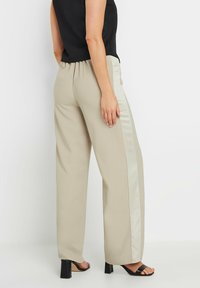 Pantalons beige à jambes larges avec une bande satinée sur le côté, offrant une texture douce et une taille élastique, portés avec des sandales à talons hauts noires.