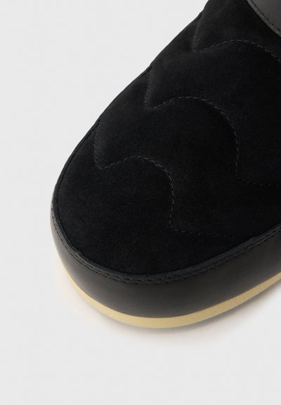 Zapatilla de ante negra con un diseño acolchado; presenta un ribete de piel suave y una suela de goma beige. Perspectiva en primer plano, destacando la textura.