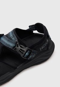 Sandal ajustável preto e azul com sola espessa e fecho de fivela, exibindo a marca "HOKA" no lado do calcanhar.