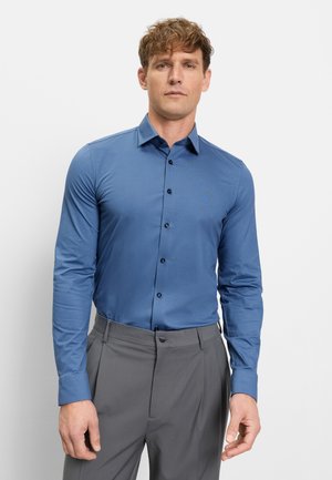 Man die een aansluitend lang blauw overhemd met knopen en een grijze broek draagt, staand tegen een eenvoudige lichte achtergrond.