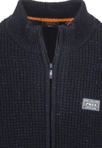 Marineblauwe gebreide zip-up cardigan met een hoge kraag, voorzien van een textuurstof en een logo-patch op de linker borst. Oranje binnenafwerking.