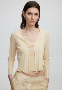Cardigan color crema con maniche lunghe, texture a costine e scollatura quadrata. Presenta una chiusura decorativa con un accento dorato.