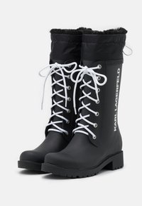 Black rubber boots with white laces and a faux fur cuff. Features a textured sole and side branding. Mid-calf length design.