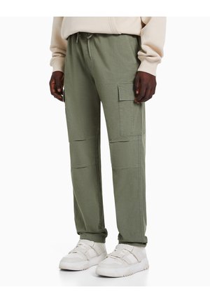 Homme portant un pantalon cargo vert olive et des baskets blanches, debout devant un fond blanc uni.
