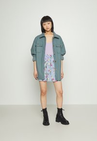 Casaco oversized em verde-água com botões frontais e dois bolsos no peito, sobre uma blusa lilás e uma saia azul com padrão floral. Botas pretas com atacadores.