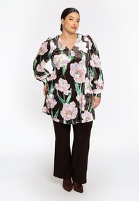 Fluwelen tuniek met een diepe V-hals, voorzien van grote roze bloemenprints op een donkerbruine achtergrond, gecombineerd met bruine flared broeken en puntige hakken.