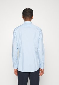 Uomo con capelli corti e intrecciati indossa una camicia a maniche lunghe a righe blu chiaro e pantaloni scuri, in piedi di spalle su sfondo bianco.