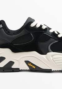 Fekete sportcipő textúrázott hálós és velúr felső résszel, fehér fűzővel, hangsúlyos Vibram logóval és egy robusztus fehér-fekete talppal.