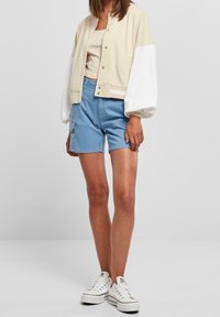 Skrácená béžovo-bílá bunda se zapínáním na patentky přes světle hnědé tričko; spárováno s modrými cargo šortkami a bílými teniskami. Hladké textury látek.
