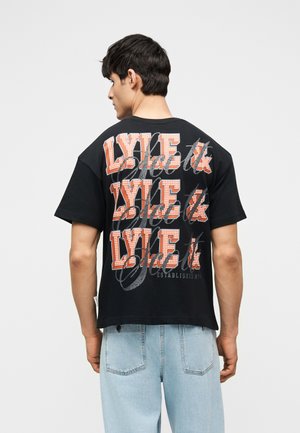 Jeune homme portant un t-shirt noir avec un grand texte orange et blanc "LYLE &" répété trois fois au dos, accompagné d'un jean bleu clair.