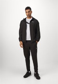 Schwarze Sportjacke mit Reißverschluss, elastischen Bündchen, kombiniert mit schwarzen Hosen und einem T-Shirt mit weißen Logo-Print. Schwarze Sneakers mit weißen Akzenten.