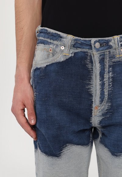 Jean en denim avec un design bicolore comportant des sections bleu foncé et gris clair, des bords effilochés et des détails de couture contrastés.