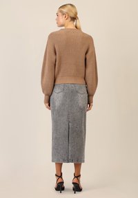 Maglione marrone lavorato a maglia con una texture a coste, abbinato a una gonna di jeans grigia con sottili decorazioni e uno spacco posteriore, indossato con tacchi neri.