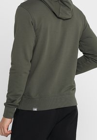 Sudadera verde oliva hecha de tela suave, con capucha ajustable, puños acanalados y una pequeña etiqueta de logo en el dobladillo. Textura suave.