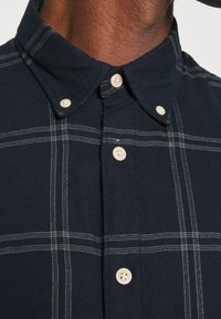Chemise bleu foncé à boutons avec des lignes à carreaux blanches et des boutons crème portée par une personne, montrant le col et la partie supérieure de la poitrine.