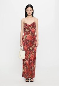 Maxi dress in a floral pattern with shades of red and green, featuring a cowl neck and spaghetti straps; paired with a white handbag.