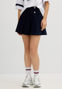 Falda plisada azul marino con una cintura alta, que presenta dos acentos de botones plateados, combinada con zapatillas plateadas y calcetines blancos con rayas rojas.