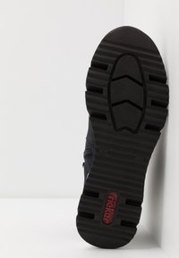Semelle de chaussure noire avec des motifs de crampons profonds et le logo "Rieker" en rouge au talon, positionnée verticalement contre un coin blanc.