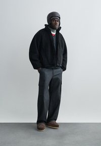 Black fleece jacket, black cargo pants, brown patterned shoes, and a gray helmet. Long sleeves, zippered closure, textured fabric.