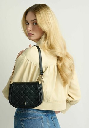 Blondynka o długich, falowanych włosach, ubrana w kremową kurtkę i niebieskie dżinsy, trzymająca czarną pikowaną torebkę na ramieniu.