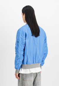 Svetlo modra bomberska jakna z gladko, sijočo površino, rebrastimi sivimi manšetami in stranskim žepom. Ima prilegajoč silhueto.