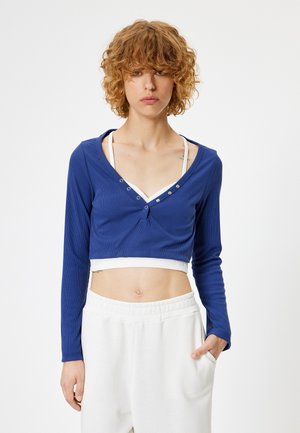 Blauw geribbeld cropped top met lange mouwen, V-hals, bijpassende witte accenten en Drukknopen. Gekocht met witte broek met elastische taille.