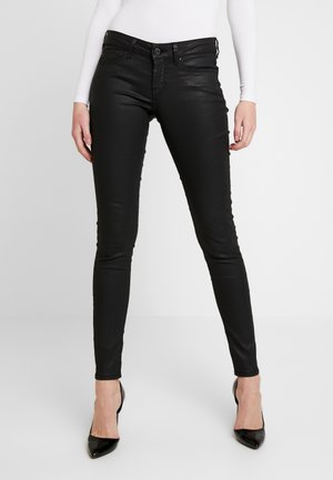 Mujer de pie con pantalones de cuero ajustados negros, tacones altos negros de punta afilada y una camiseta blanca de manga larga, sobre un fondo liso.