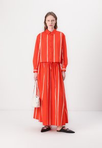 Set di camicia e gonna a righe rosse. La camicia ha il colletto e i bottoni; la gonna è lunga e con vita regolabile. Abbinata a scarpe nere e una borsa bianca.