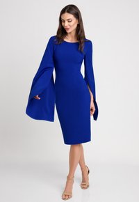 Niebieska dopasowana sukienka z długimi, rozszerzanymi rękawami i bocznymi rozcięciami. Wykonana z gładkiego materiału, do kolan, z okrągłym dekoltem. Beżowe sandały na wysokim obcasie.