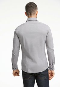 Maglietta grigia a maniche lunghe con colletto, dotata di polsini con bottoni e tessuto testurizzato. Indossata con jeans scuri, evidenziando una silhouette aderente.