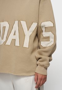 Beżowy sweter z dużymi białymi aplikacjami w kształcie liter "DAYS" oraz cyfrą "5". Posiada luźny krój i ściągacze przy mankietach. Teksturowana tkanina.