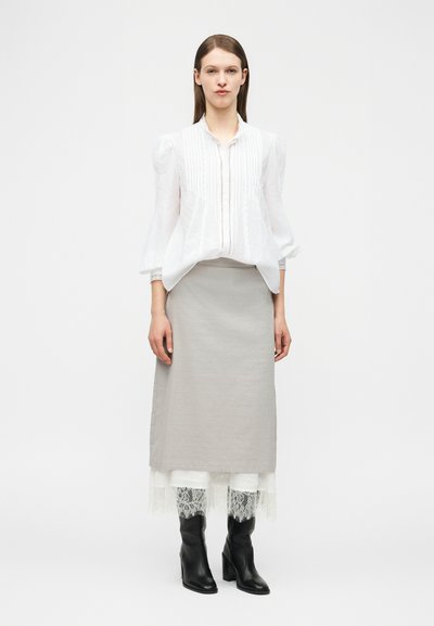 Γυναίκα όρθια φορώντας λευκό πλισέ πουκάμισο, μπεζ μεσοφόρι με λευκή δαντέλα στο τελείωμα και μαύρα μπότες με τακούνι μπροστά σε απλό φόντο.