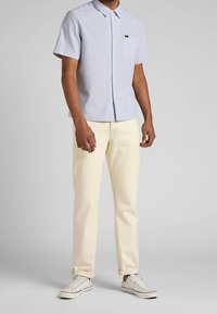 Camicia azzurra a maniche corte con colletto button-down, taschino sul petto, abbinata a pantaloni color crema e scarpe da ginnastica bianche.