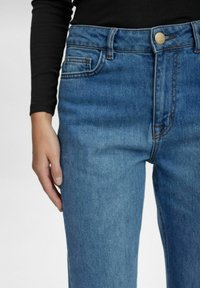 Srednje visoki modri traperji z značilnim ravnim krojem, subtilnim zbledenjem, svetlim šivanjem in kovinskim gumbom za zapiranje v pasu.