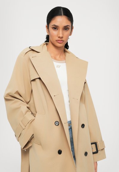 Μπεζ παλτό trench με διπλό κούμπωμα, μεγάλες πέτα και μαύρα κουμπιά. Διαθέτει πλάγιες τσέπες και ζώνη στη μέση.