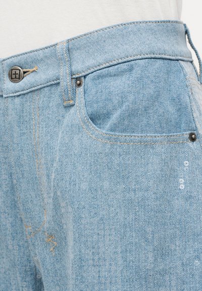 Φωτεινό μπλε τζιν σε κοντινό πλάνο που δείχνει τη μέση, την μπροστινή τσέπη, το μεταλλικό κουμπί, τα τρουκ και τις λεπτομερείς ραφές.
