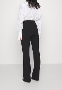 Kobieta w czarnych, wysokich, szerokich spodniach i białej koszuli z długim rękawem, stojąca na tle jasnego, jednolitego tła.