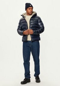 Giacca puffer blu navy con cappuccio e design trapuntato; foderata in beige; indossata sopra un maglione chiaro e jeans blu; abbinata a stivali neri e un berretto.